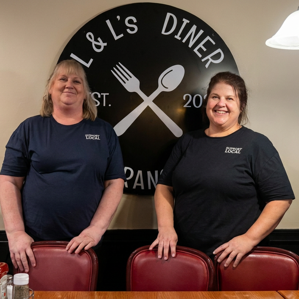 Two smiling restaurant staff members stand behind red dining chairs beneath an L & L’s Diner sign.