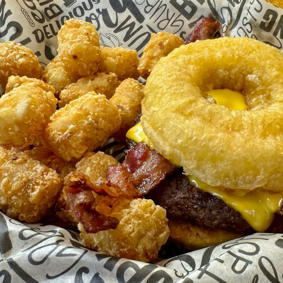 Cheeseburger with bacon, fried egg, and a ring-shaped bun served alongside crispy golden potato bites on printed paper.