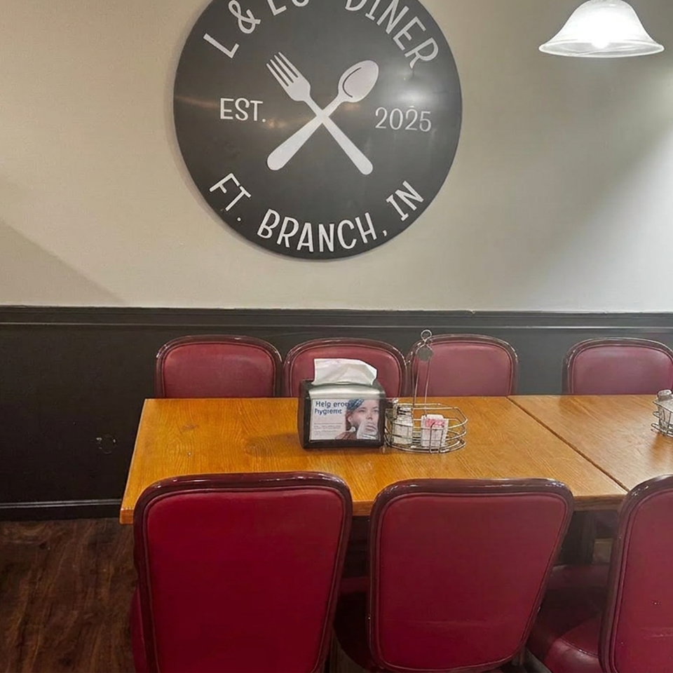 Restaurant dining area with wooden tables, burgundy chairs, pendant lighting, and a wall sign reading L&L Diner, Est. 2025, Ft. Branch, IN.